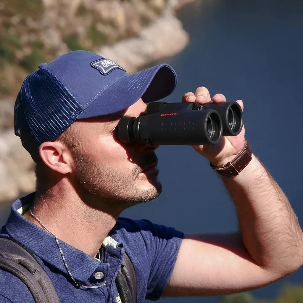 Persona utilizando binoculares Tasco 10x42 negros durante una excursión al aire libre, con fondo de paisaje montañoso y lago.